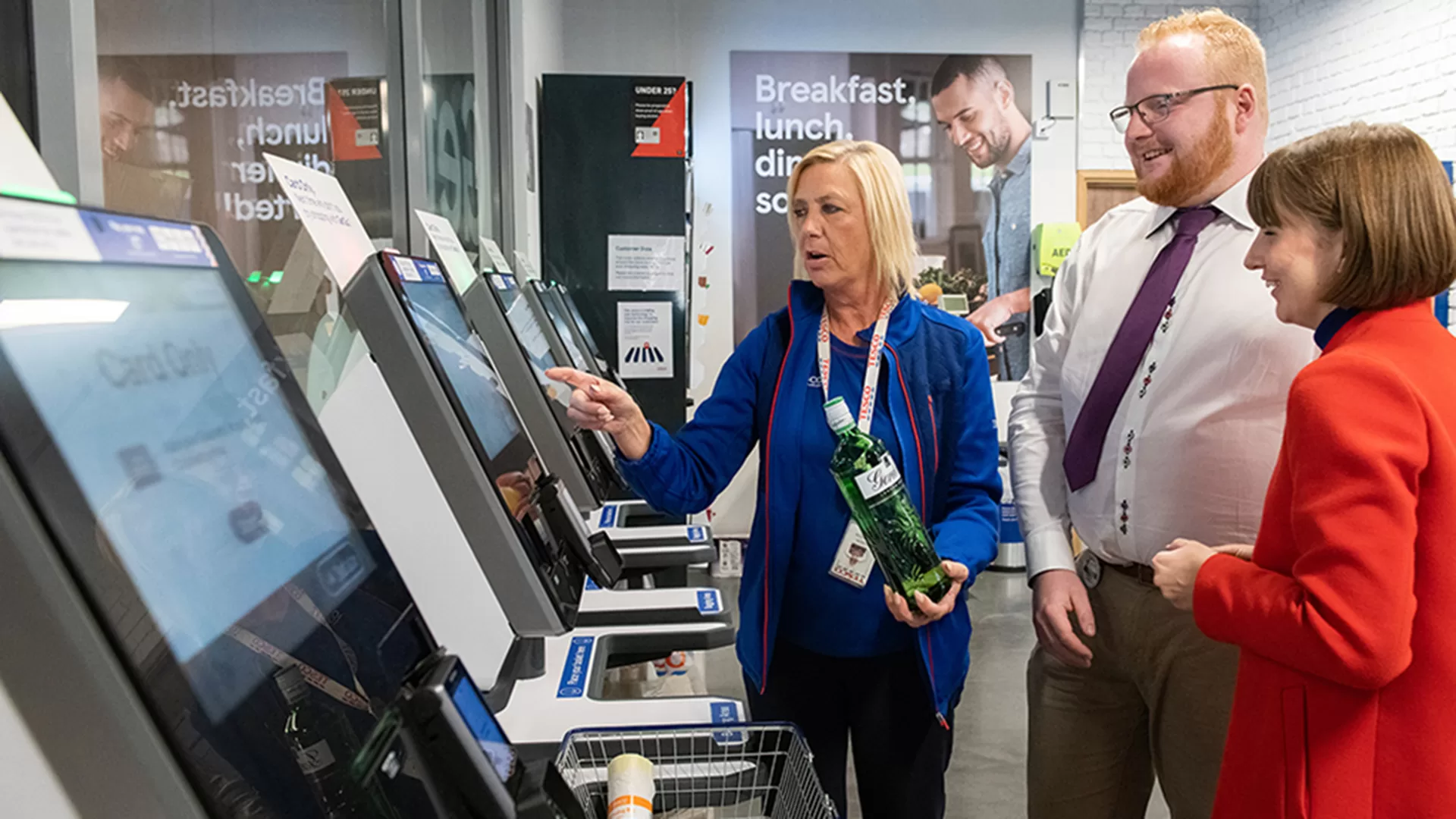 Tesco sales assistant helping two people with a self service till
